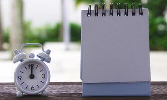 Alarm clock pointing at 12 o'clock with blank notepad. Blurred park background. Copy space photo