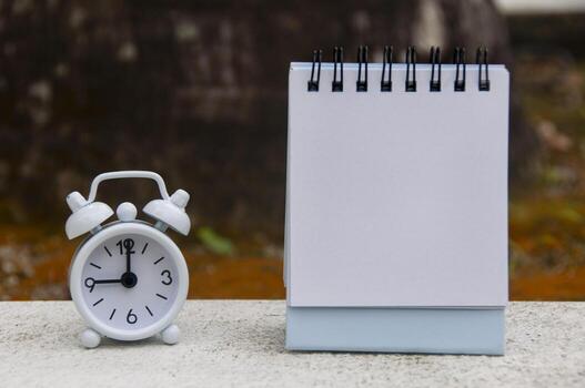 Alarm clock pointing at 9 o'clock with blank notepad. Blurred park background. Copy space concept photo