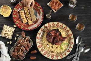 mano femenina elige baklava turco en el plato, mesa para el saludo de ramazán con dátiles, arroz árabe, pollo asado, nueces y té foto