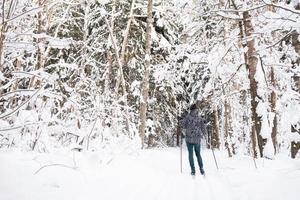 Skier in hat with pompom with ski poles in his hands with his back against the background of a snowy forest. Cross-country skiing in winter forest, outdoor sports, healthy lifestyle. photo