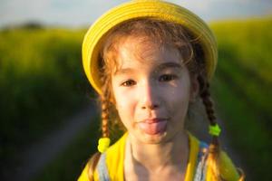 A cheerful girl in a yellow hat in a summer field hooligan shows the tongue and teases. Carelessness, joy, sunny weather, holidays. Lifestyle, kind and funny face, close-up portrait photo