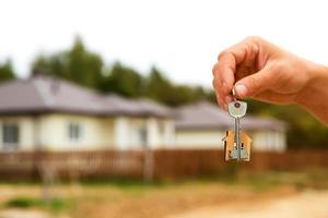 Hand with a key and a wooden key ring-house. Background of fence and cottage. Building, project, moving to a new home, mortgage, rent and purchase real estate. To open the door. Copy space photo