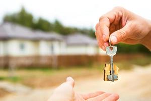 Hand with a key and a wooden key ring-house. Background of fence and cottage. Building, project, moving to a new home, mortgage, rent and purchase real estate. To open the door. Copy space photo