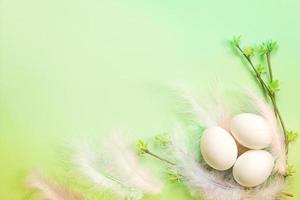 White Easter eggs in a delicate nest of colored feathers with unfurled leaf buds on the branches. Spring, a religious holiday, the birth of life. Copy space. Pale green background photo