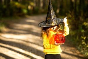 A girl in a witch's hat, in a yellow sweater and with a pumpkin a lantern and a bucket for sweets in autumn forest. Halloween Holiday. Portrait, close-up photo