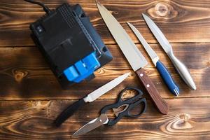 Sharpening a knife on an electric sharpener at home. Layout of knives and scissors with a sharpening machine on a wooden table. Top view, flat lay. photo