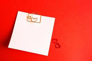 Square sheet for notes with a paper clip in shape of a letter and a heart. Decor of couples in love with hearts on a red background. Valentine's Day, message, greeting, declaration of love. Copy space photo