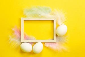 Easter white frame on a bright yellow background of chicken eggs and colored delicate feathers. Spring, religious holiday, Easter decoration, greeting, copy space, mock up photo
