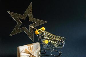 Shopping cart with a gold transparent star with sequins in the shape of a Christmas tree on a black background. Black Friday, buying gifts for the new year. Space for text photo