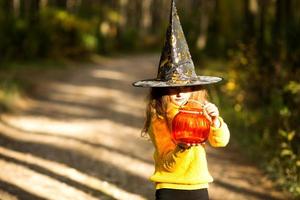 A girl in a witch's hat, in a yellow sweater and with a pumpkin a lantern and a bucket for sweets in autumn forest. Halloween Holiday. Portrait, close-up photo