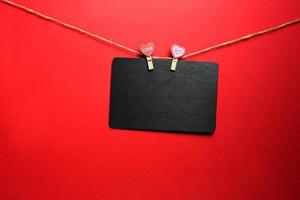 A brown wooden writing Board with copyspace hangs on a rope with two clothespins with hearts and the inscription Love. Valentine's day, mock up for lovers. Red background, frame photo