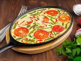 Oval cast iron pan with delicious hot omelette with tomatoes and capsicum. Brown wood texture background, close-up, side view. photo