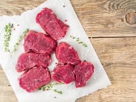 Pieces of raw meat. Raw beef with spices and thyme on white parchment paper on wooden rough rustic background, top view, close up, copy space. photo