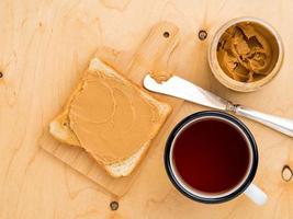 tostadas con mantequilla de maní, cuchillo para untar en un sándwich, una taza de té sobre un fondo de madera beige foto