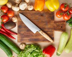 Vegetables around wooden cutting board with knife on kitchen table. Top view. photo