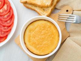 ramekin redondo blanco con tortilla de huevos y leche al horno, con una costra, sobre una mesa de madera blanca, vista superior. foto