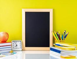 Chalkboard, apple and school supplies on white table by the yellow wall. Side view, empty space. Back to school concept. photo
