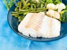Fillet of boiled sea fish cod with broccoli, green beans and cauliflower on a blue plate, side view, close. Healthy diet dish for proper nutrition photo
