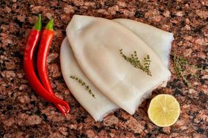 Three carcasses of fresh raw squid with lemon, pepper, thyme on a marble stone dark table. photo