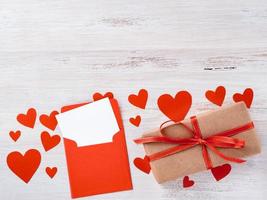 symbol of Valentine's day - gift box in kraft brown paper with red ribbon, envelope with a clean blank sheet of paper on white background with lots of red hearts, side view