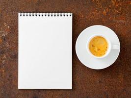 blank sheet of notebook with a spiral and cup of coffee on rusted old iron table, top view.
