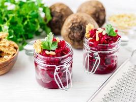 Fresh salad of grated boiled beetroot in glass jars, white wooden background, side view. photo
