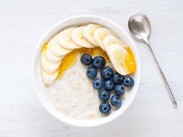 Oatmeal with bananas, blueberries, chia, jam, honey on white wooden background. Healthy breakfast. Top view, photo