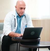 Male doctor is using notebook computer in hospital living room. Professional medical person in white coat and stethoscope sits relaxing looking at computer screen for diagnosis, healthcare information photo