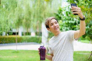 apuesto joven deportista asiático posando y sonriendo a la cámara mientras bebe agua, hombre escuchando música en el smartphone mientras practica el entrenamiento de carrera. feliz atleta asiático sonriendo. foto