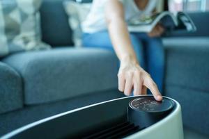 Happy Asian young woman turning on high efficiency air purifier while staying and reading a book in the living room. Woman using air purifier in her house. photo