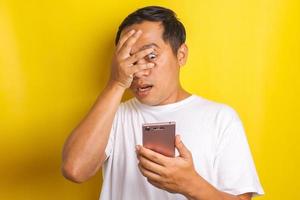 Close-up portrait of shocked, scared asian man closing one eye while looking at cell phone isolated on yellow background photo
