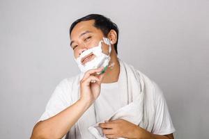 Handsome asian man shaving. Handsome asian man shaving his face and looking at camera while standing on grey background photo