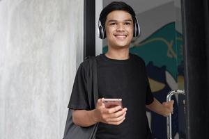 Happy young asian man wearing wireless headphones smiling while opening glass door photo
