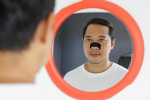 cara de hombre asiático con parche para el cuidado de la piel en la nariz. retrato de hombre joven mirando al espejo con una tira de limpieza de poros en una piel limpia y fresca. foto