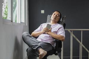 joven asiático sentado relajándose mientras disfruta de la música a través del teléfono móvil en casa foto