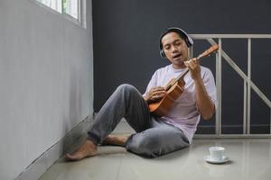 un apuesto joven asiático sentado relajándose en el suelo tocando el ukelele foto