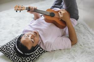 joven asiático relajándose, acostado tocando el ukelele mientras canta solo en casa foto