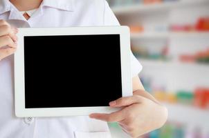 farmacéutico con bata blanca que muestra una tableta digital en blanco con estantes de medicamentos en la farmacia foto