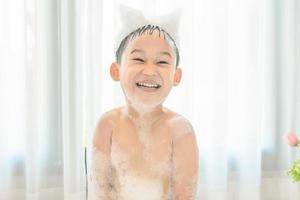 smiling happy asian child boy is playing with white foam in tub bath at home. photo