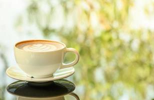 Latte art coffee cup on glass table. Blurred leaf reflection background. photo