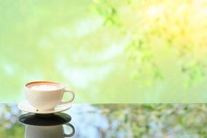 Latte art coffee cup on glass table. Blurred leaf reflection background. photo