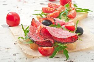 Sandwich with sausage, olive, tomato and arugula on the old wooden background photo