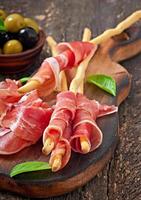 Grissini bread sticks with ham, olives, basil on old wooden background photo