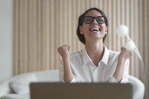 Excited italian lady clenching fists with joy while sitting in front of laptop at workplace