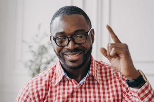 un tutor de inglés masculino de etnia africana sonriente con gafas levanta la mano y señala con el dedo índice hacia arriba foto