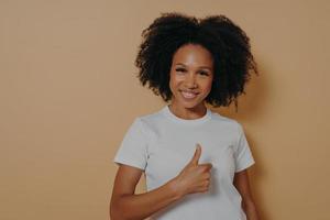 Cheerful positive young african woman saying well done, showing thumb up while standing in studio photo