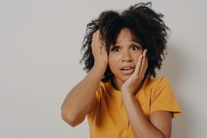 Shocked and nervous dark skinned female student holding hands on head and gasping with worried face photo