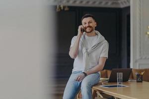 Happy bearded male freelancer works in home office room dressed in casual t shirt and jeans has telephone conversation discusses working issues has cheerful expression works remotely from home photo