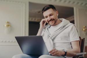 hombre alegre freelancer ajustando, auricular y mirando la computadora portátil mientras habla por video chat con colegas foto