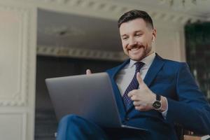 Successful bearded male entrepreneur dressed in formal suit uses modern laptop computer and earbuds for making web conference chatting keeps thumbs raised makes like gesture wears blue formal suit
